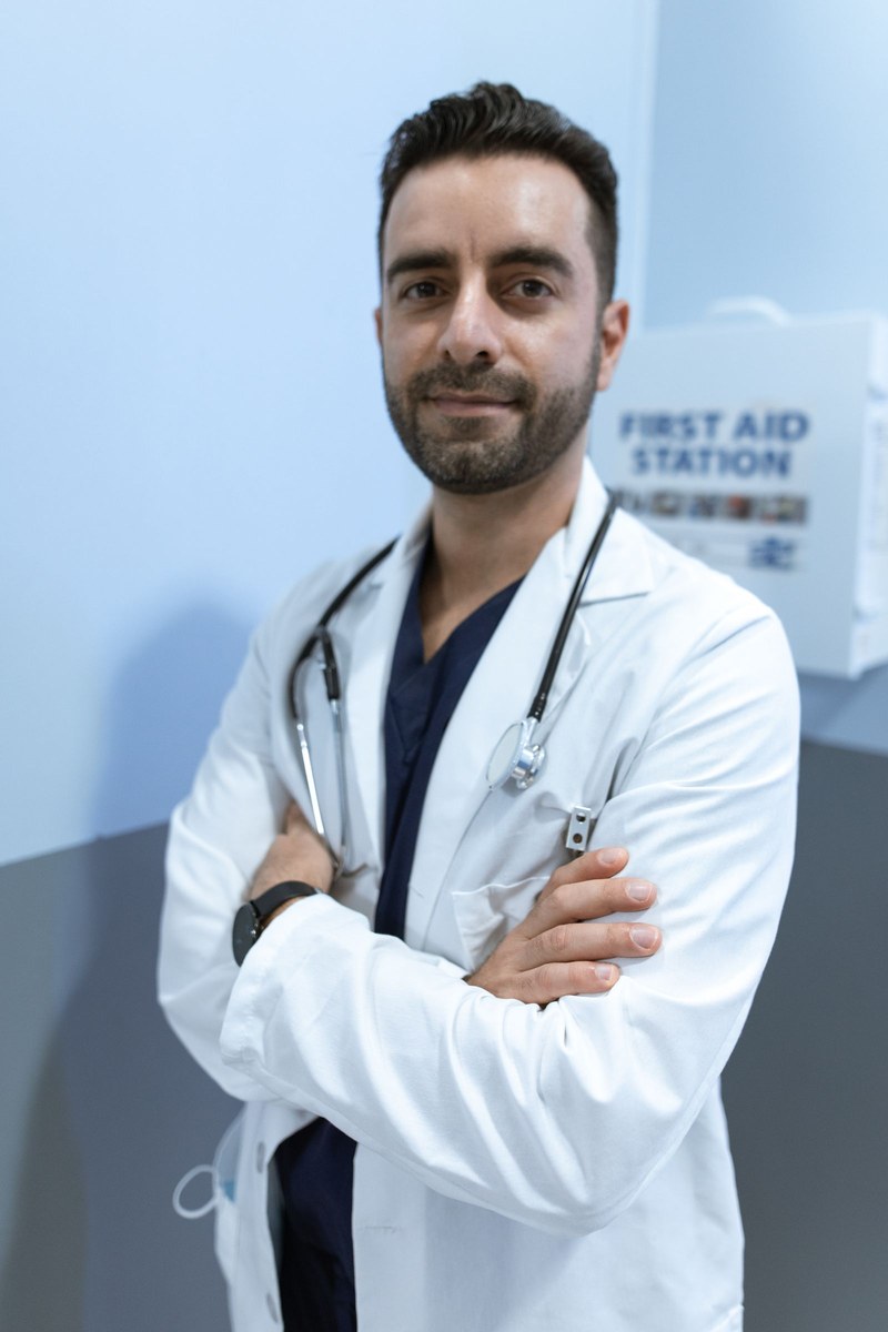Male doctor with a dark beard, white coat, and black stethoscope standing arms crossed by a first-aid station