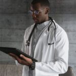 Male doctor in a white coat and gold glasses reviewing a black tablet against a grey clinic wall
