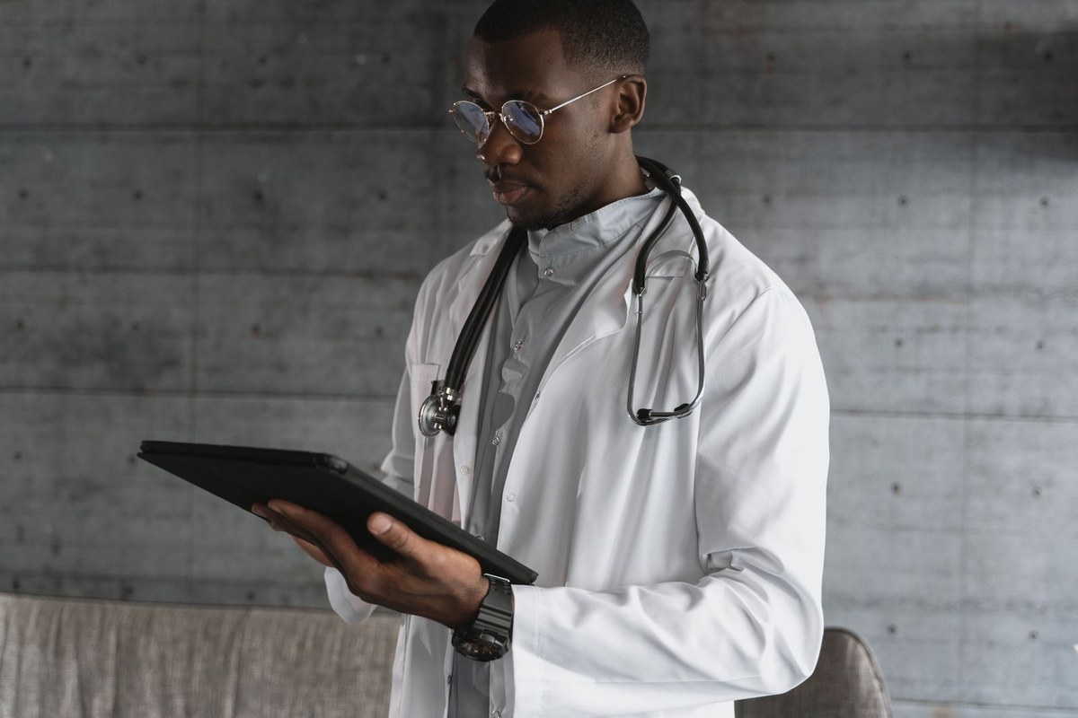 Male doctor in a white coat and gold glasses reviewing a black tablet against a grey clinic wall