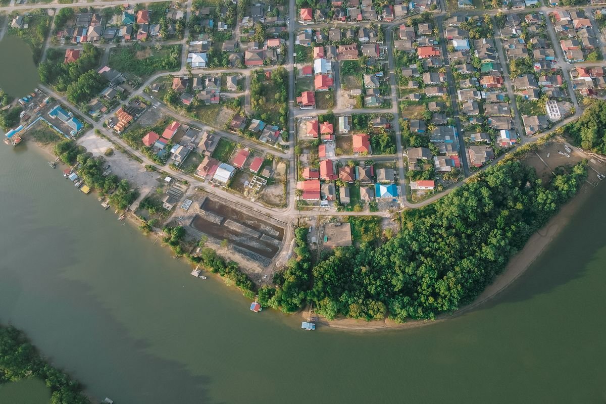 Aerial view of a low-rise housing area with red and grey roofs and narrow roads beside a river lined with trees